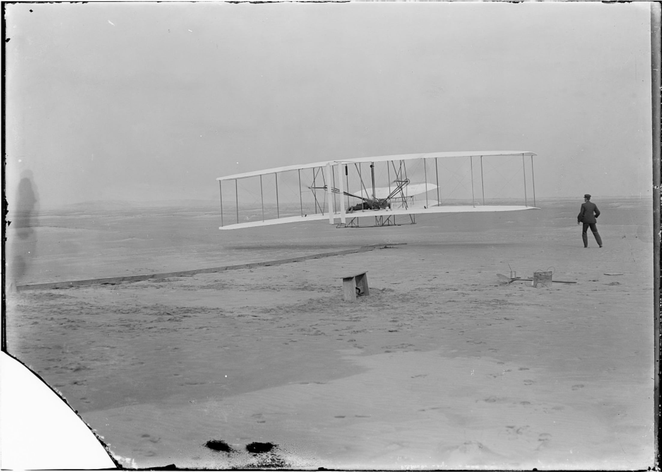 This is the most famous of the Wright brothers photographs, depicting the 1903 flyer a few seconds after it took off in the first powered heavier-than-air flight. The landscape is a flat sand bar. Orville Wright is at the controls. Wilbur is in the photograph, having trotted beside the flyer as it rolled down the launching rail. He has just let go of the wing and the flyer is about three feet off the ground.