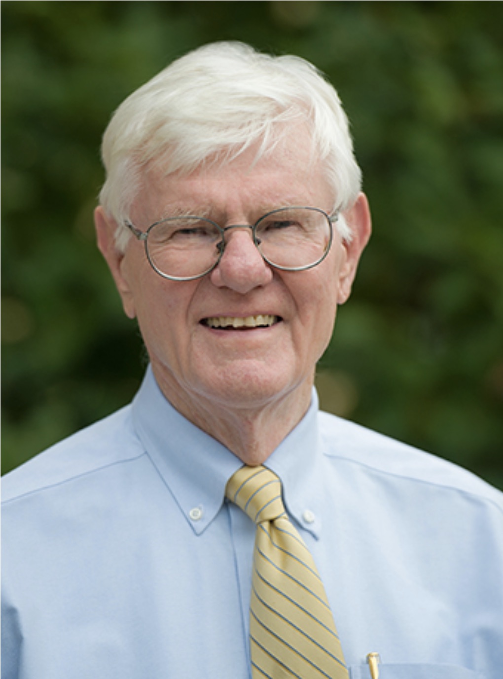 Portrait of an older man with white hair and glasses, wearing a light blue shirt and a yellow striped tie, outdoors.
