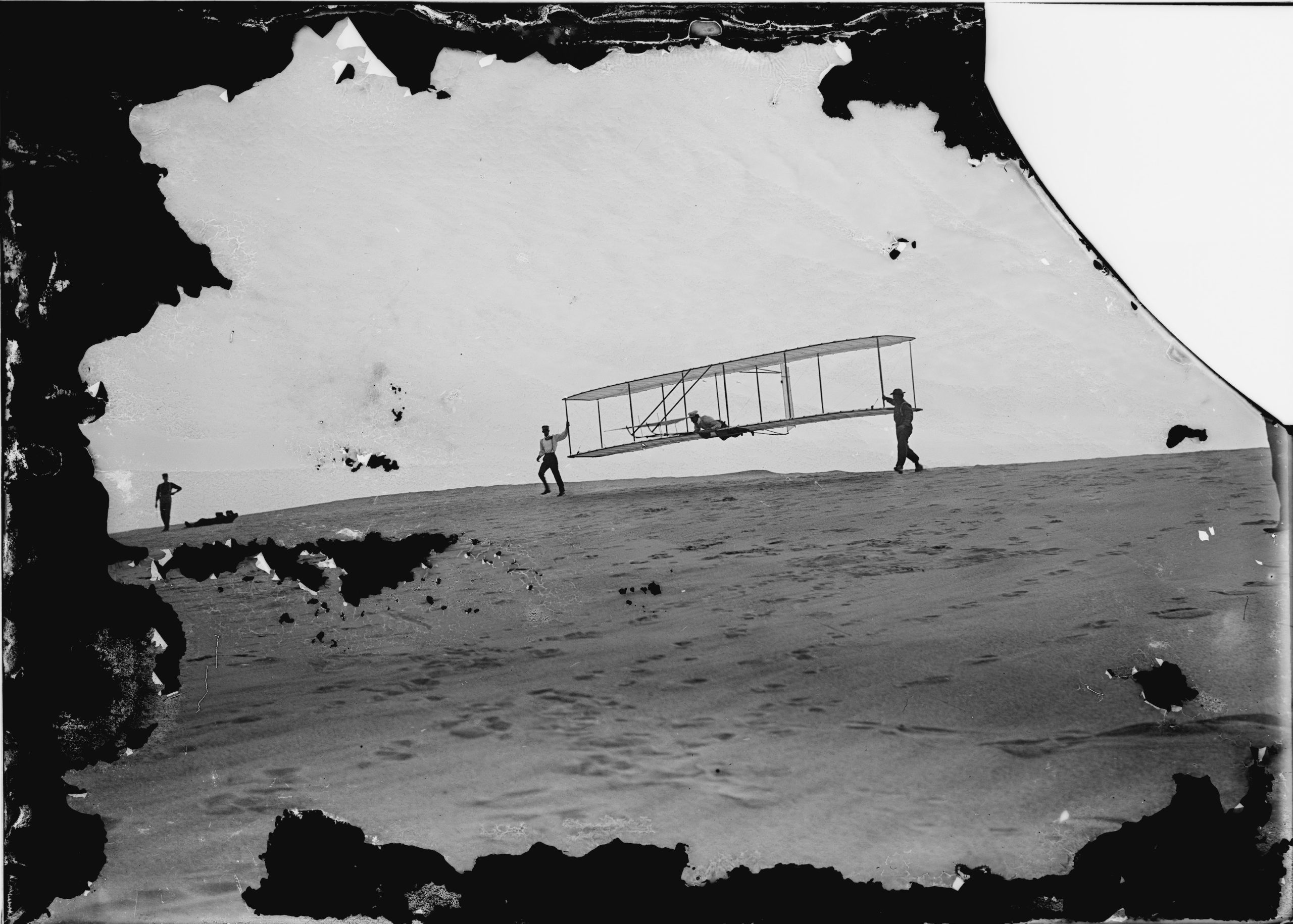 This is a Wright brothers photograph of the 1902 glider being launched from a sand dune. Two men are holding up the glider while the operator lays on the lower wing.