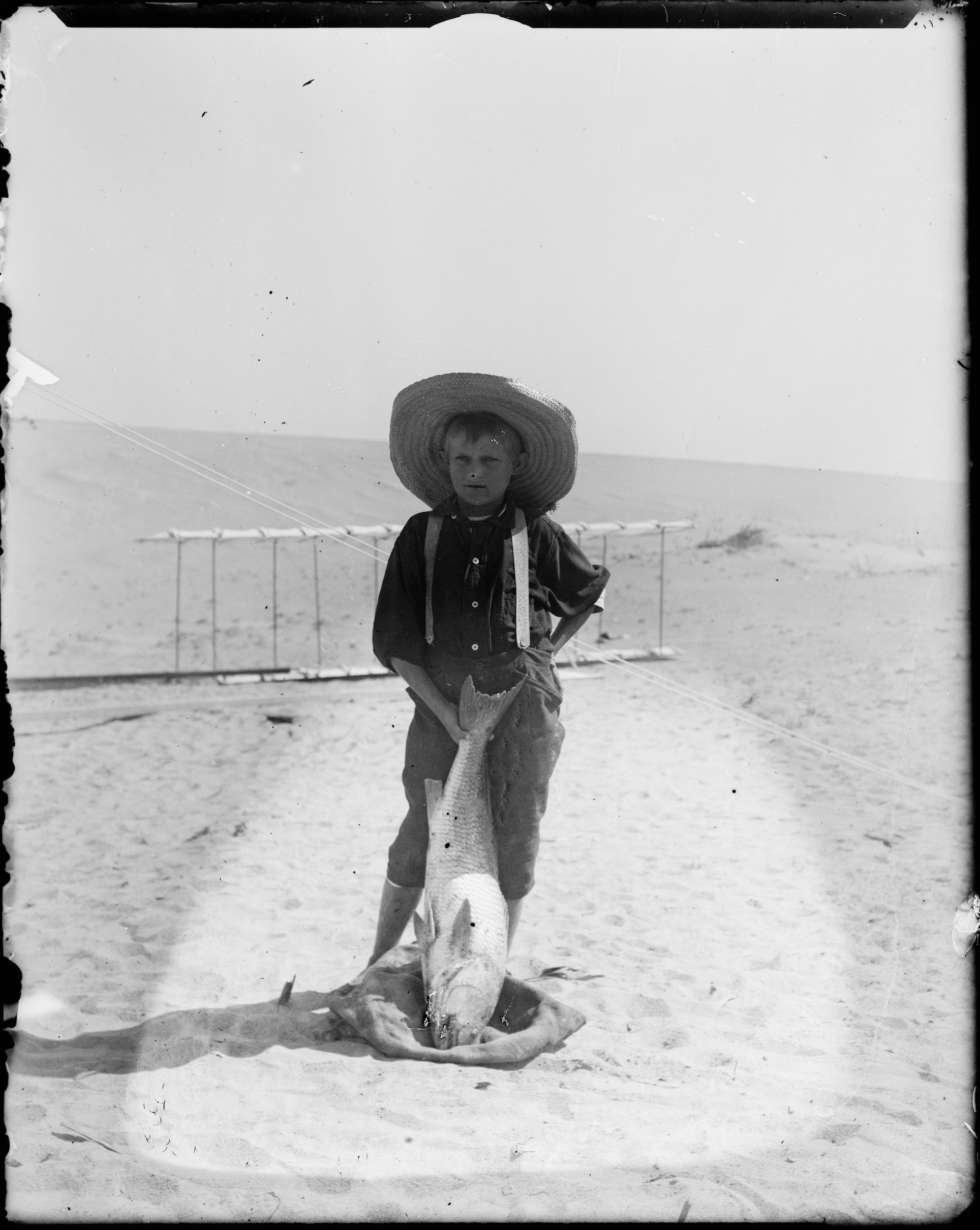 This is a photograph taken by the Wright brothers of Tom Tate, a young boy, posing with a large fish in front of the Wright's 1900 glider. Tom is wearing a straw hat and suspenders.