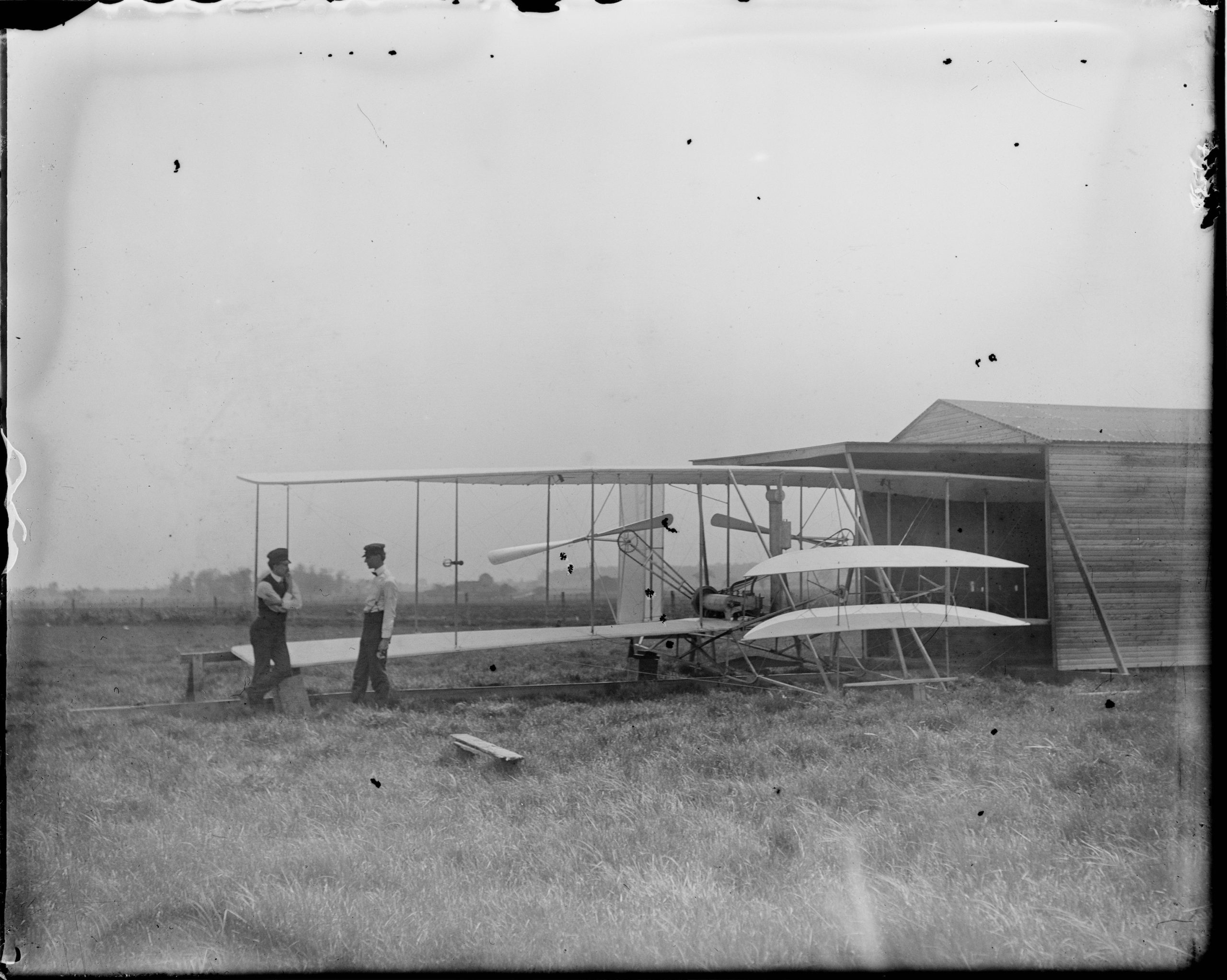 This is a photograph of the Wright brothers with their 1904 flyer at Huffman Prairie. Their testing ground is an open, treeless expanse of grass. At right is the storage shed they built to hold their flying machine.